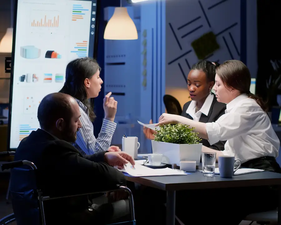 Four colleagues around a meeting table reviewing printed charts, with a dashboard on the screen behind them